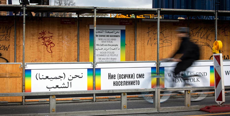 Ein Bauzaun ist mit Plakaten beklebt. Auf den Plakaten steht in verschiedenen Sprachen „Wir (alle) sind das Volk“.