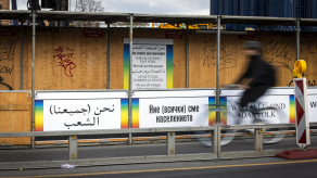 Bauzaun mit Plakaten. Darauf steht in verschiedenen Sprachen „Wir (alle) sind das Volk“.